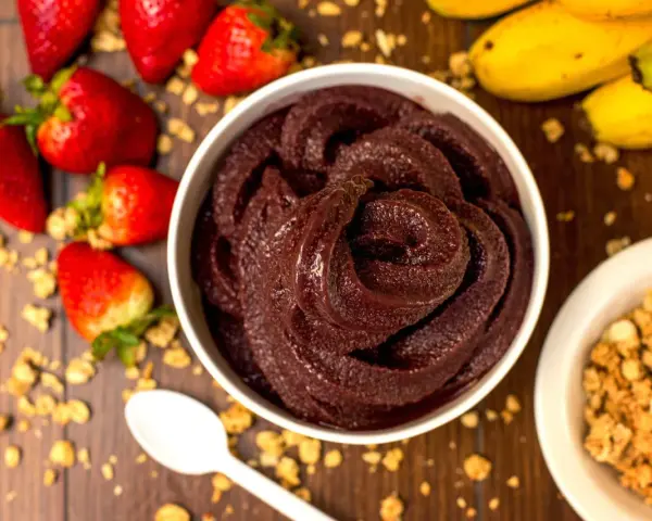 Un bol de sorbet **açaí** onctueux, entouré de fraises, bananes et granola. Une cuillère blanche est posée à côté.
