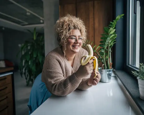 Une femme souriante avec des cheveux bouclés tient une banane, malgré une possible allergie à la banane.