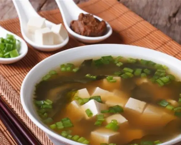 Un bol de soupe miso avec des cubes de tofu, des algues et des oignons verts ciselés. Trois cuillères présentent des ingrédients : oignons verts, tofu et pâte miso.