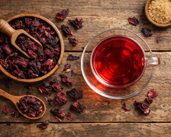 Infusion hibiscus bienfaits : une tasse de thé rouge vif à côté d'un bol de fleurs d'hibiscus séchées et d'une cuillère en bois.