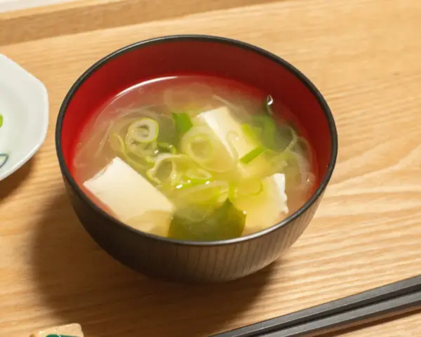 Un bol de soupe japonaise, avec du tofu et des oignons verts, servi avec des baguettes sur un plateau en bois.