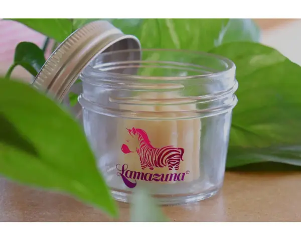 Petit pot en verre ouvert, révélant un baume solide. Le logo Lamazuna, un zèbre rose, orne le verre. Un soin naturel à base de beurre de cacao.