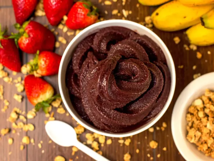 Un bol de sorbet **açaí** onctueux, entouré de fraises, bananes et granola. Une cuillère blanche est posée à côté.