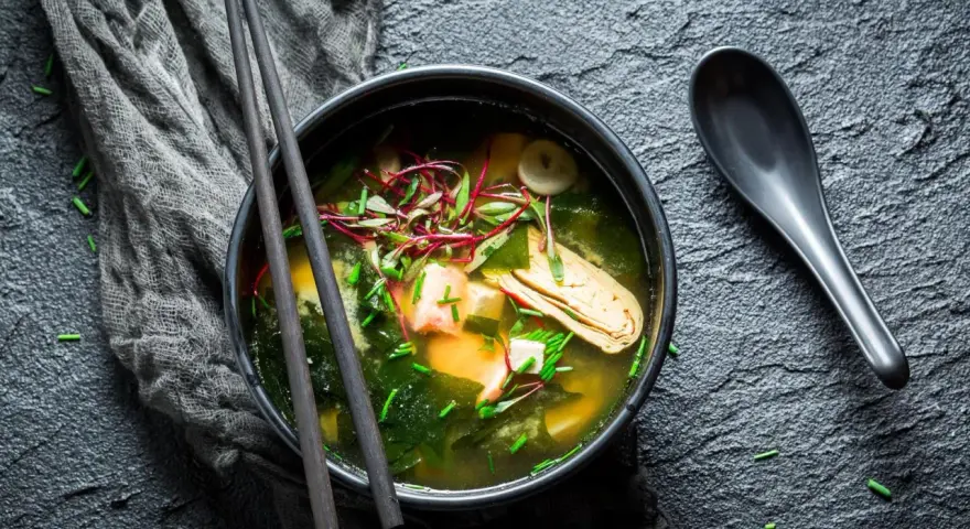 Un bol de bouillon miso fumant, garni de tofu, d'algues et de pousses rouges, prêt à être dégusté avec des baguettes.