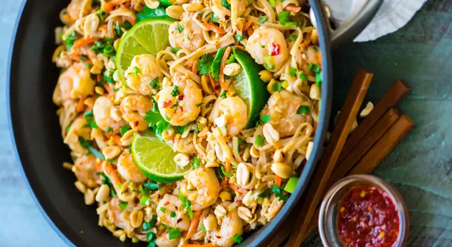 Un plat de recette pad thai crevette appétissant dans un wok, garni de crevettes, de cacahuètes, de coriandre et de quartiers de citron vert.