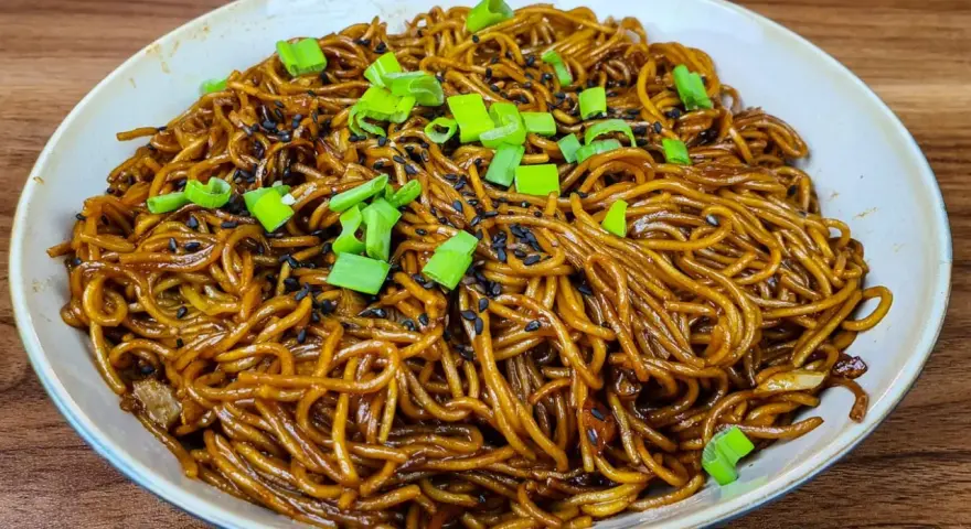 Un plat de nouilles savoureuses, enrobées d'une sauce brune appétissante, garnies de graines de sésame noir et de ciboule fraîche.