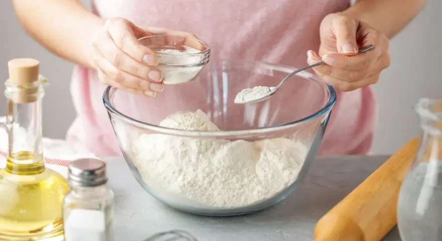 Une personne ajoute une poudre blanche à de la farine dans un bol. Une cuillère et un petit bol sont utilisés. C'est une idée pour par quoi remplacer la levure chimique.