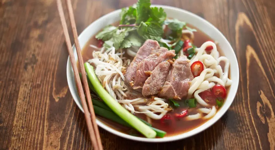 Un bol de soupe nouille de riz avec du bœuf, des germes de soja, des oignons verts et du piment rouge, prêt à être dégusté avec des baguettes.