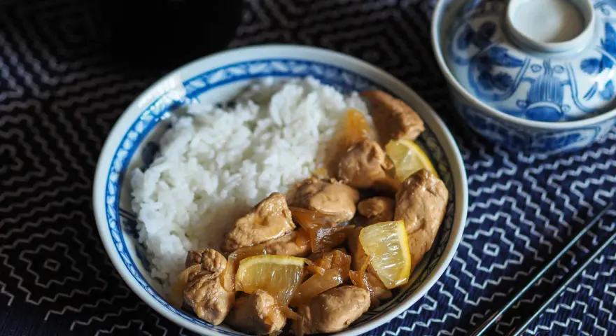 Un plat de riz blanc avec du poulet mariné sauce soja, garni de quartiers de citron. Un bol décoré et des baguettes complètent la scène.