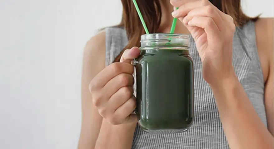 Une femme boit un smoothie vert à la spiruline, un allié pour la santé et la beauté des cheveux.