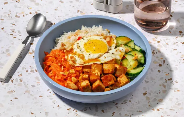 Un bol de bibimbap tofu avec riz, carottes râpées, concombre, œuf au plat et cacahuètes concassées.