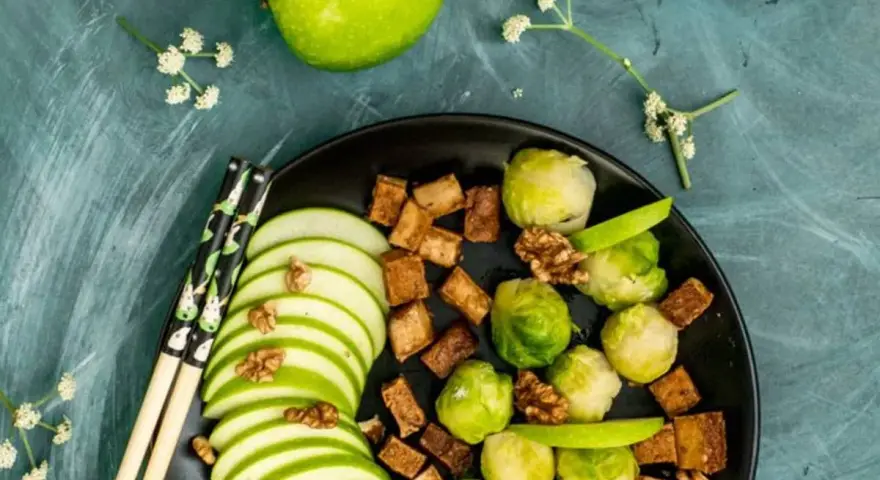 Salade colorée avec des tranches de pomme verte, des choux de Bruxelles, des dés de tofu croustillant et des noix, le tout arrosé d'une vinaigrette miso.