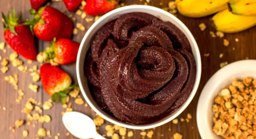 Un bol de sorbet **açaí** onctueux, entouré de fraises, bananes et granola. Une cuillère blanche est posée à côté.