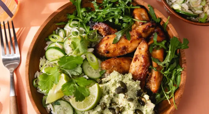 Un bol de riz au miso avec poulet glacé, concombre, coriandre, roquette et une purée verte parsemée de graines.