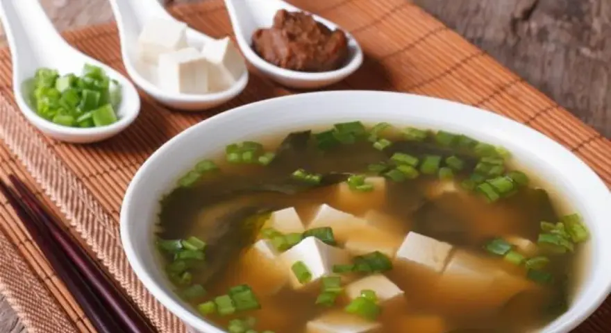 Un bol de soupe miso avec des cubes de tofu, des algues et des oignons verts ciselés. Trois cuillères présentent des ingrédients : oignons verts, tofu et pâte miso.