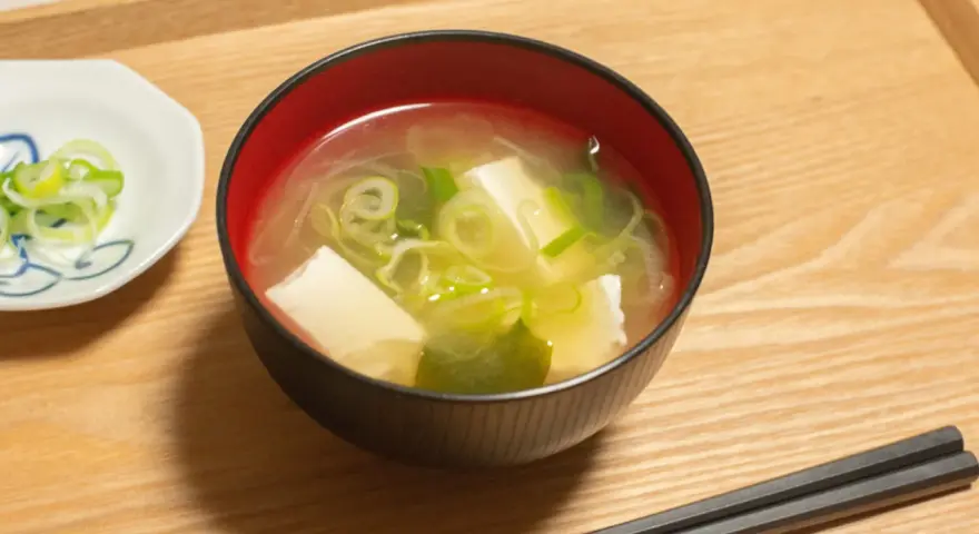 Un bol de soupe japonaise, avec du tofu et des oignons verts, servi avec des baguettes sur un plateau en bois.