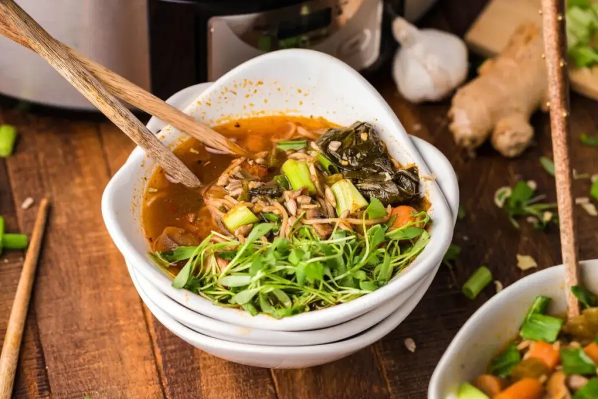 Un bol de ramen veggie fumant, garni de pousses fra&icirc;ches, de c&eacute;leri et de l&eacute;gumes verts. Des baguettes en bois reposent sur le bord.