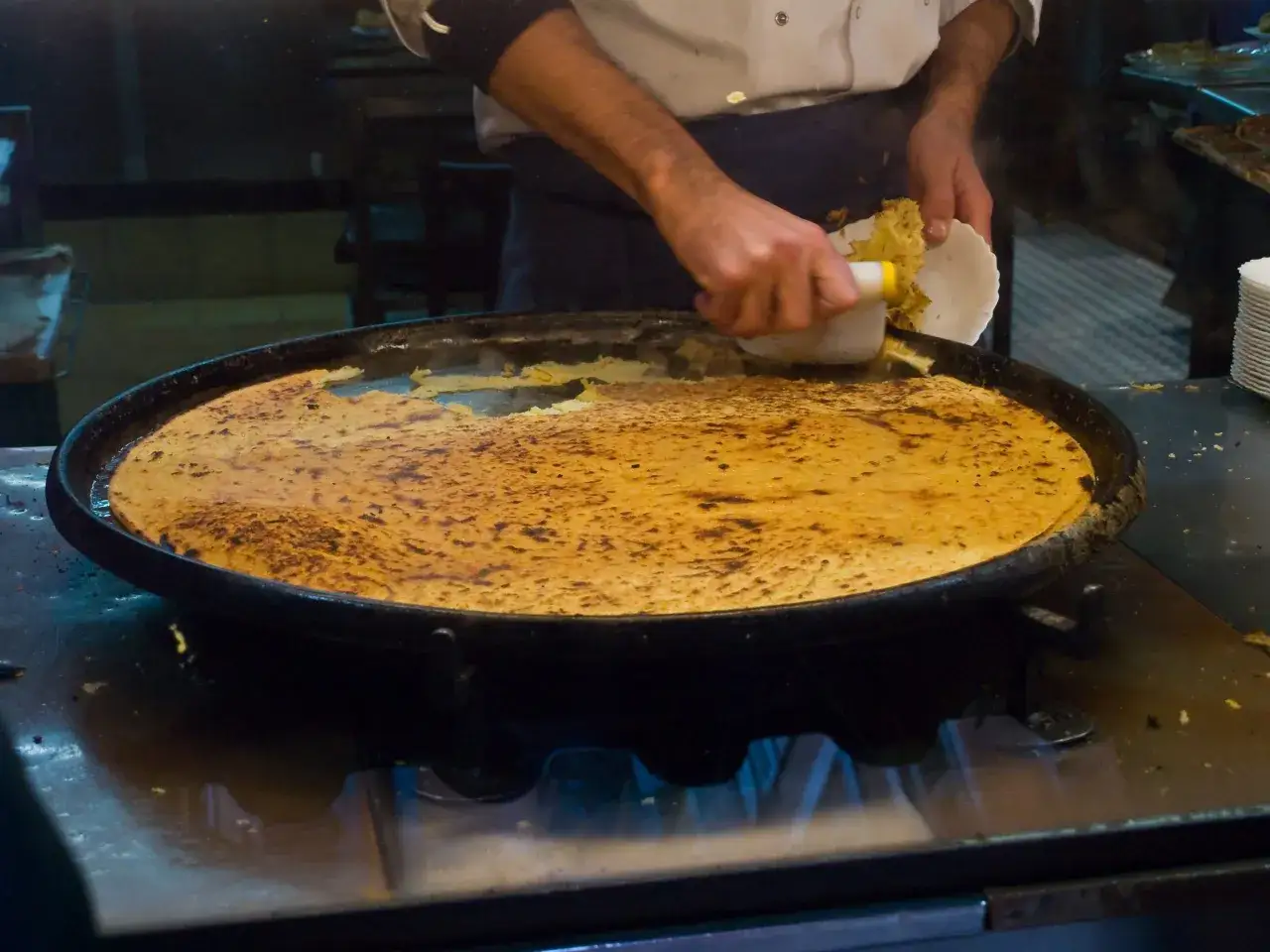 Un cuisinier pr&eacute;pare une galette dor&eacute;e &agrave; la po&ecirc;le, &agrave; base de farine de pois chiche, qui cr&eacute;pite sur le feu.