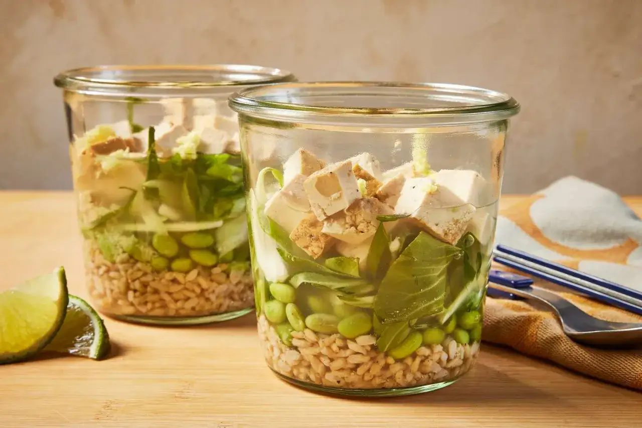 Deux bocaux de soupe japonaise avec riz, edamame, tofu et l&eacute;gumes verts. Des quartiers de citron vert sont &agrave; c&ocirc;t&eacute;.