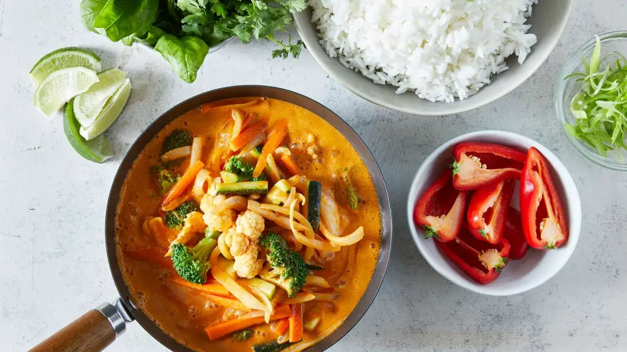 Un plat de riz au curry v&eacute;g&eacute;tarien, avec du brocoli, du chou-fleur, des carottes et des courgettes dans une sauce cr&eacute;meuse. Servi avec du riz blanc, des quartiers de citron vert et des poivrons rouges coup&eacute;s.