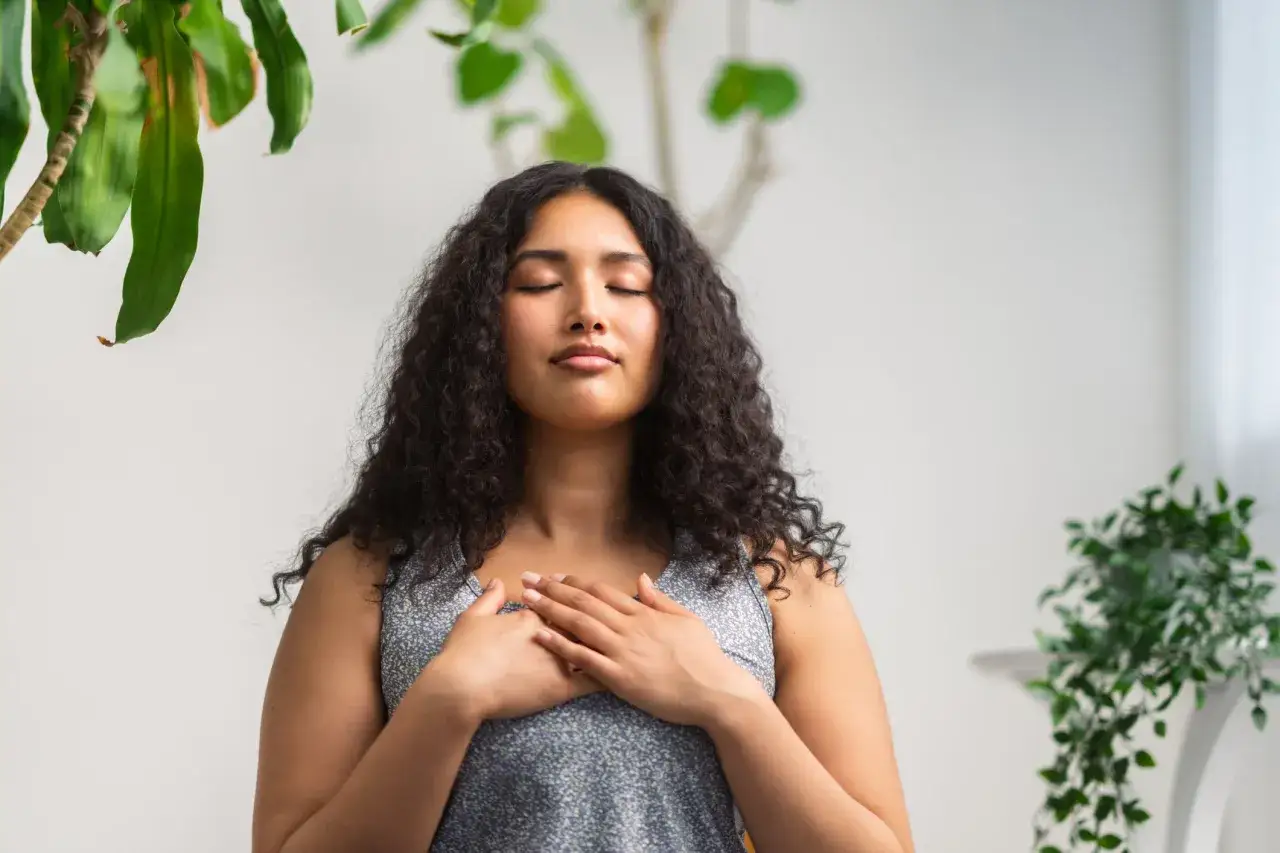 Jeune femme aux yeux fermés, mains sur le cœur, entourée de plantes. Une scène paisible pour apprendre comment réduire le stress naturellement.