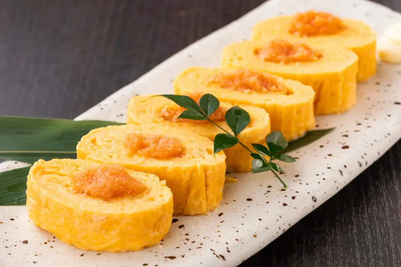 Rouleaux d'oeuf japonais, dor&eacute;s et moelleux, garnis d'une touche orang&eacute;e, pr&eacute;sent&eacute;s sur une assiette d&eacute;corative avec une feuille verte.