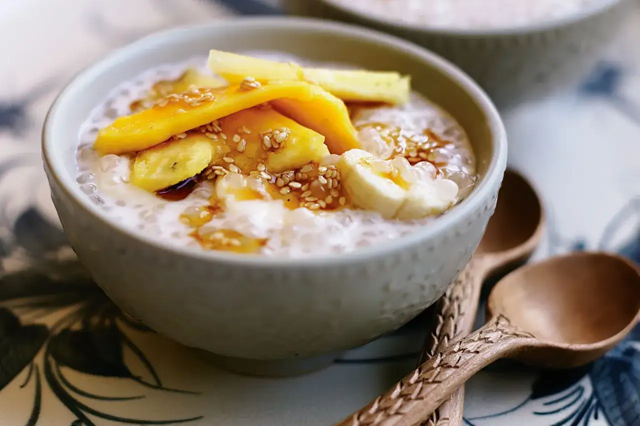 Un bol de banane au lait de coco, garni de fruits et de graines de sésame, prêt à être dégusté avec des cuillères en bois.
