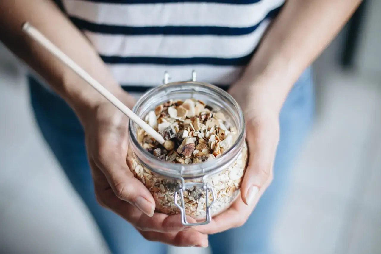 Mains de personne tenant un bocal en verre rempli de muesli maison, avec des flocons d'avoine, des noix et des fruits secs.