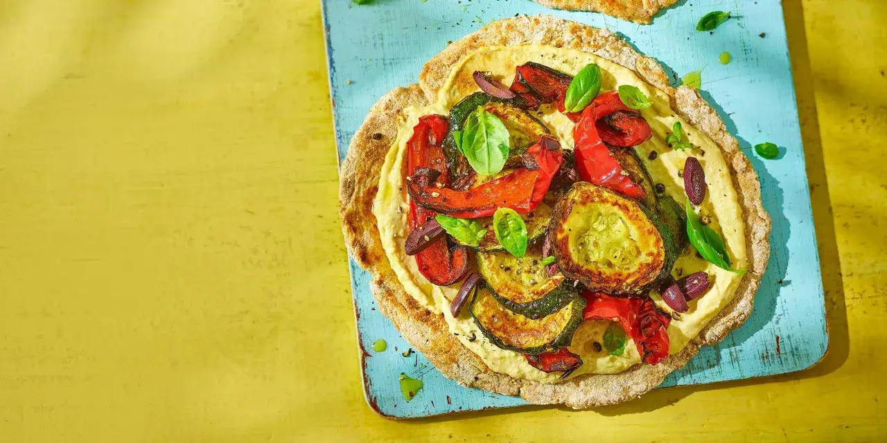 D&eacute;licieux d&eacute;jeuner au bureau : galette garnie de houmous, l&eacute;gumes grill&eacute;s (courgettes, poivrons) et olives, sur fond jaune vif.